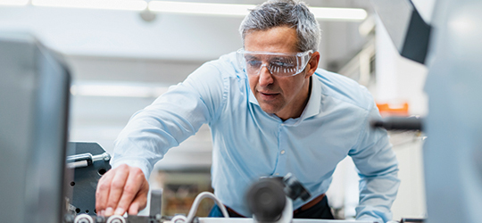 A man in business casual clothes, wearing eye protectors, checking large printer.