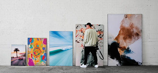 person in street clothes stands with back facing camera, looking at canvas prints lined up along a wall