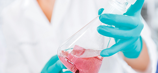 a scientist in a white lab coat wears blue latex gloves while holding a beaker containing fluid and a textile